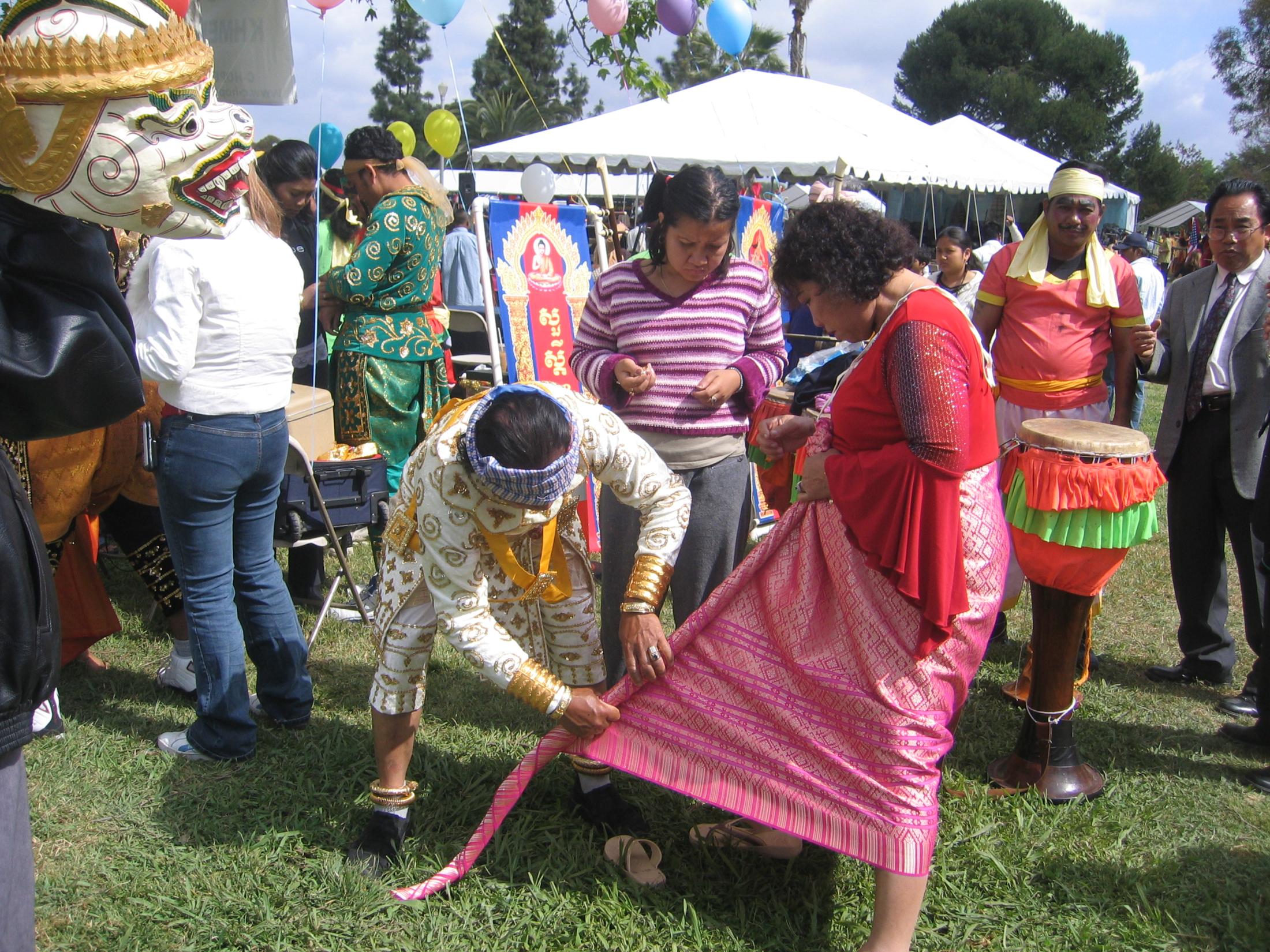 Performers help each other get dressed for their performances at Cambodian New Year in Long Beach. The woman is being helped with her kbun. An 8’ length of silk is wrapped around her waist toward the front. The length of cloth will be twisted and pulled to her back between her legs. It is held in place with a belt.
