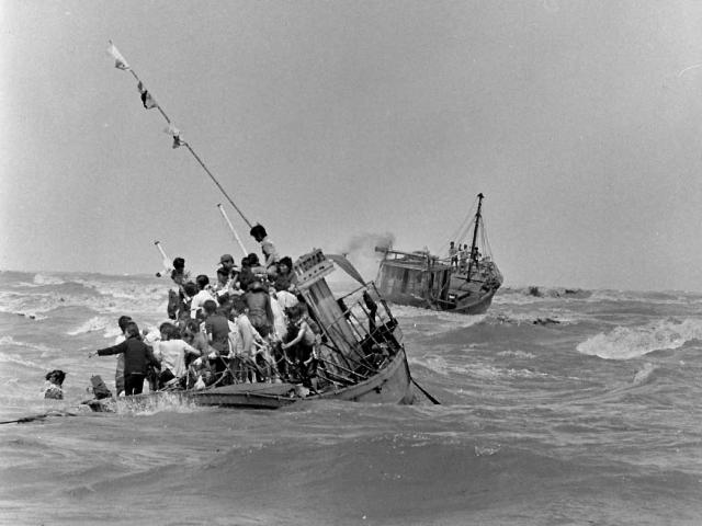 A boat crowded with Vietnamese men, women and children in heavy seas; ahead of it and travelling in the same direction is another boat with a small number of people standing on the deck.