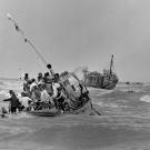 A boat crowded with Vietnamese men, women and children in heavy seas; ahead of it and travelling in the same direction is another boat with a small number of people standing on the deck.